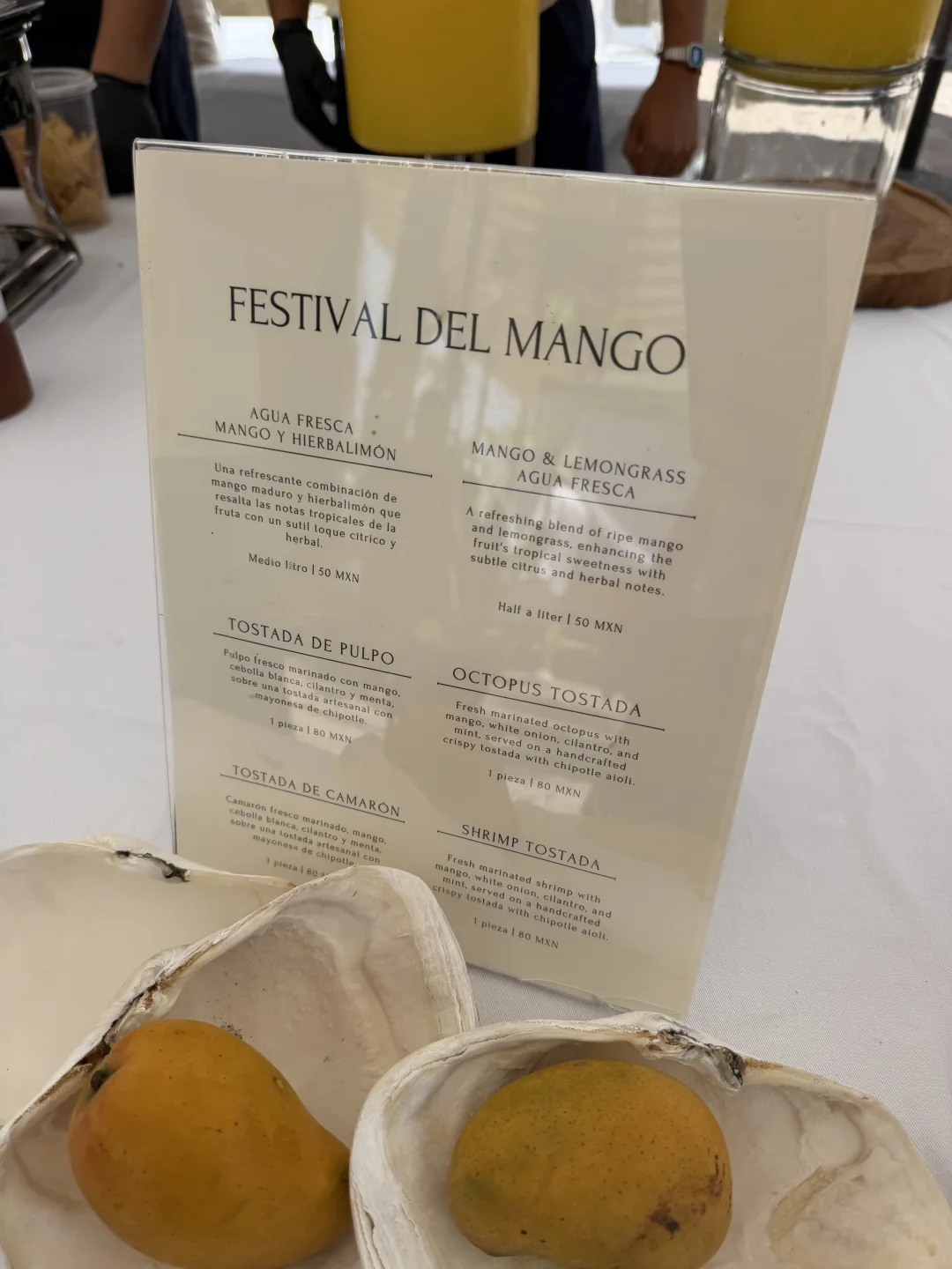 Festival menu beside fresh mangoes, advertising mango agua fresca and seafood tostadas